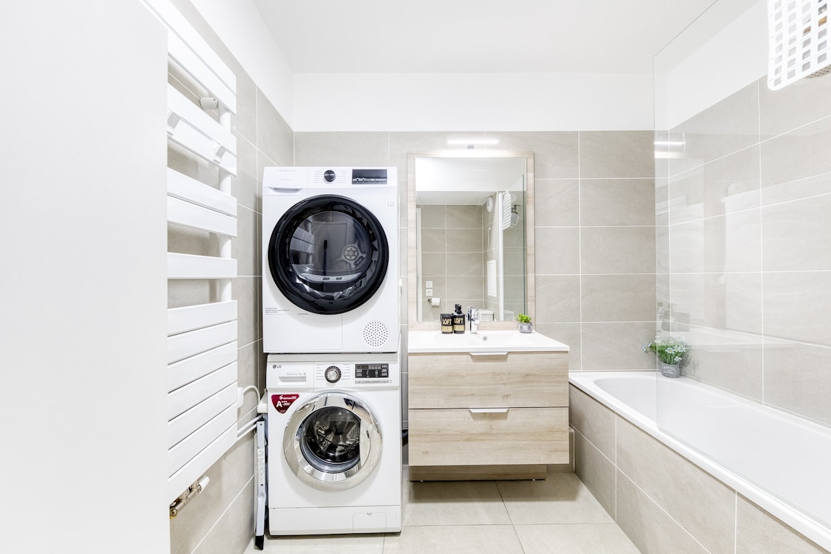 Salle de bain — Salle de bain moderne avec lave-linge et sèche-linge empilés, carrelage beige clair, baignoire et vasque bois.