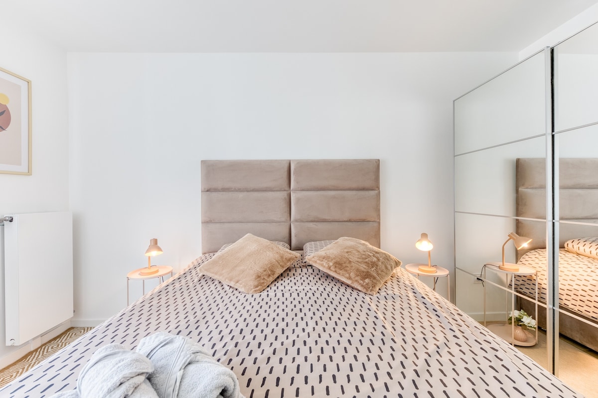 Chambres — Lit beige spacieux, miroir placard, lumière chaleureuse