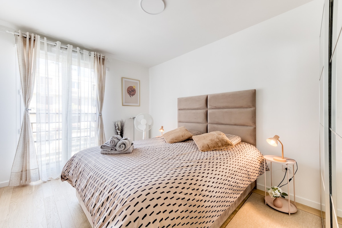 Chambres — Lit spacieux, tête de lit capitonnée beige, lumière naturelle généreuse